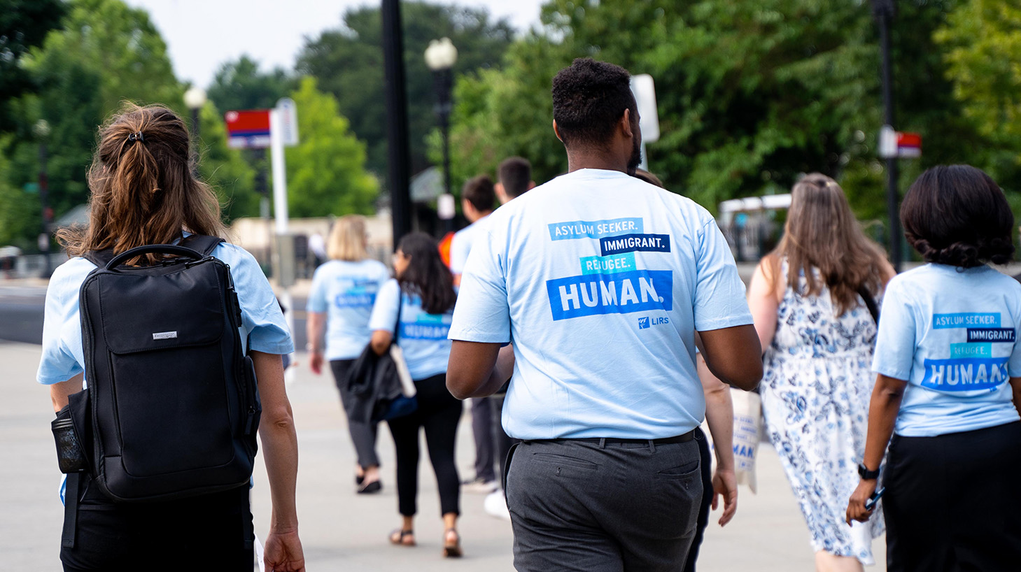 Backs of a group of people walking in Global Refuge T-shirts that say Asylum Seeker. Immigrant. Refugee. Human.